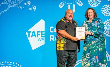 Two people standing in front of a blue backdrop with white decorative patterns. The backdrop has the text 'TAFE WA' and 'Central Regional' on it. One person is holding a certificate, and both are dressed in colorful clothing.
