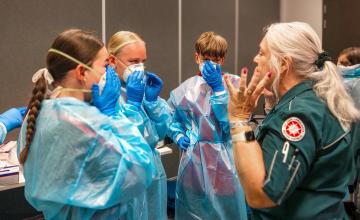Group of individuals in blue protective gowns, gloves, and face masks gathered around an older person in a dark green uniform with a red and white emblem on the sleeve, in a room with gray walls and a table with items.