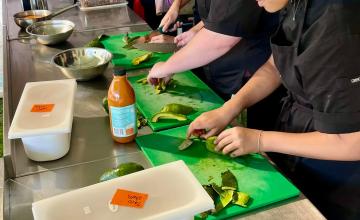 A group of people in a kitchen setting, likely participating in a cooking class or workshop. They are wearing black aprons and working on green cutting boards placed on a stainless steel countertop. Various ingredients and utensils are visible, including avocados being cut, bottles of sauce, bowls, and plastic containers with orange labels. The background has text that reads 'KIDS IN THE KITCHEN.
