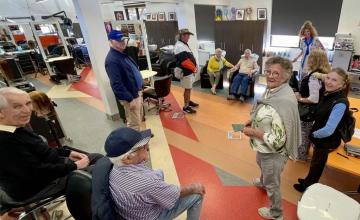 A group of people gathered in a well-lit room with colorful flooring, chairs, and tables. Some are seated while others are standing. The walls are decorated with framed pictures and artwork.